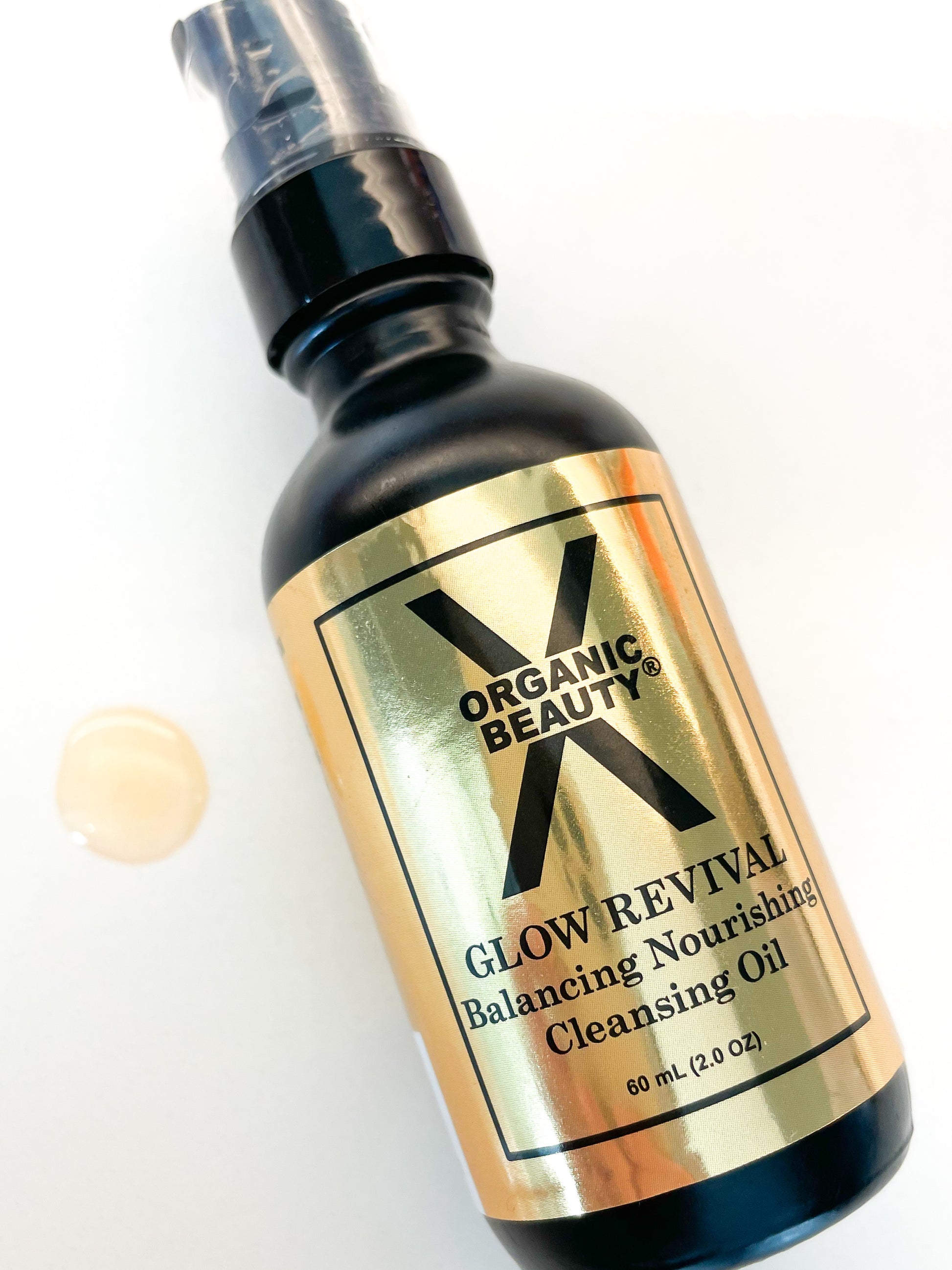 Organic Glow Revival Cleansing Oil bottle on a white background with a orange/gold drop of oil next to it