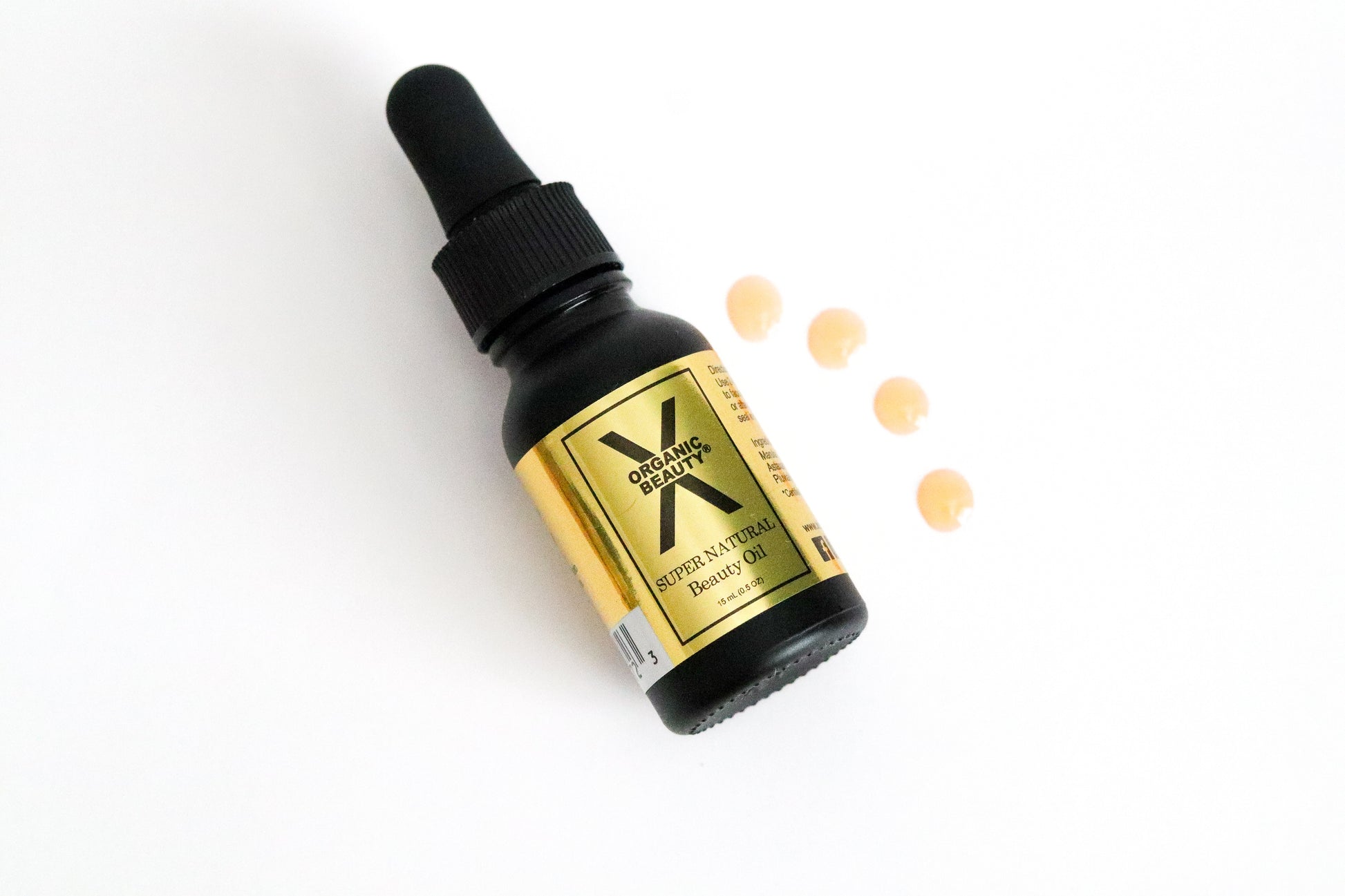 A half ounce bottle of Super Natural Beauty Oil on it's side with four drops of the orange liquid beside it on a white background