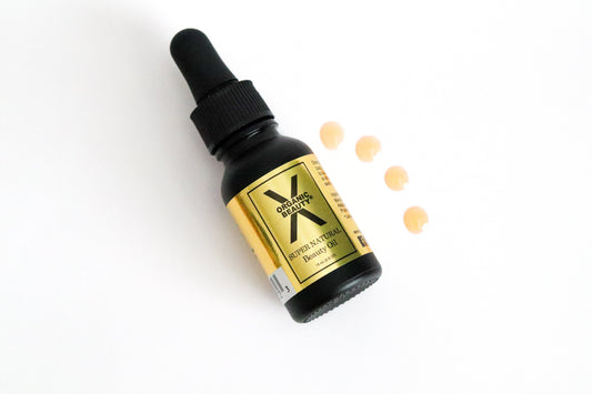 A half ounce bottle of Super Natural Beauty Oil on it's side with four drops of the orange liquid beside it on a white background