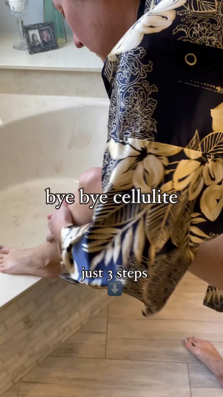 A person in a bathroom wearing a black and gold floral robe, leaning over a bathtub with text overlays that read 'bye bye cellulite' in 'just 3 steps' showing how to use Super Natural Beauty Oil with a fascia roller to remove cellulite
