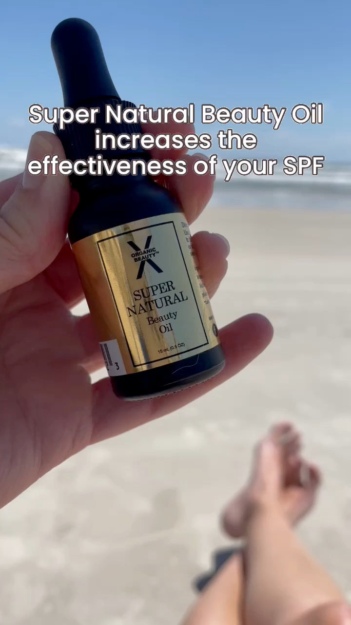 A hand holds a small bottle of Super Natural Beauty Oil against a beach background with text overlay reading 'Super Natural Beauty Oil increases the effectiveness of your SPF'.