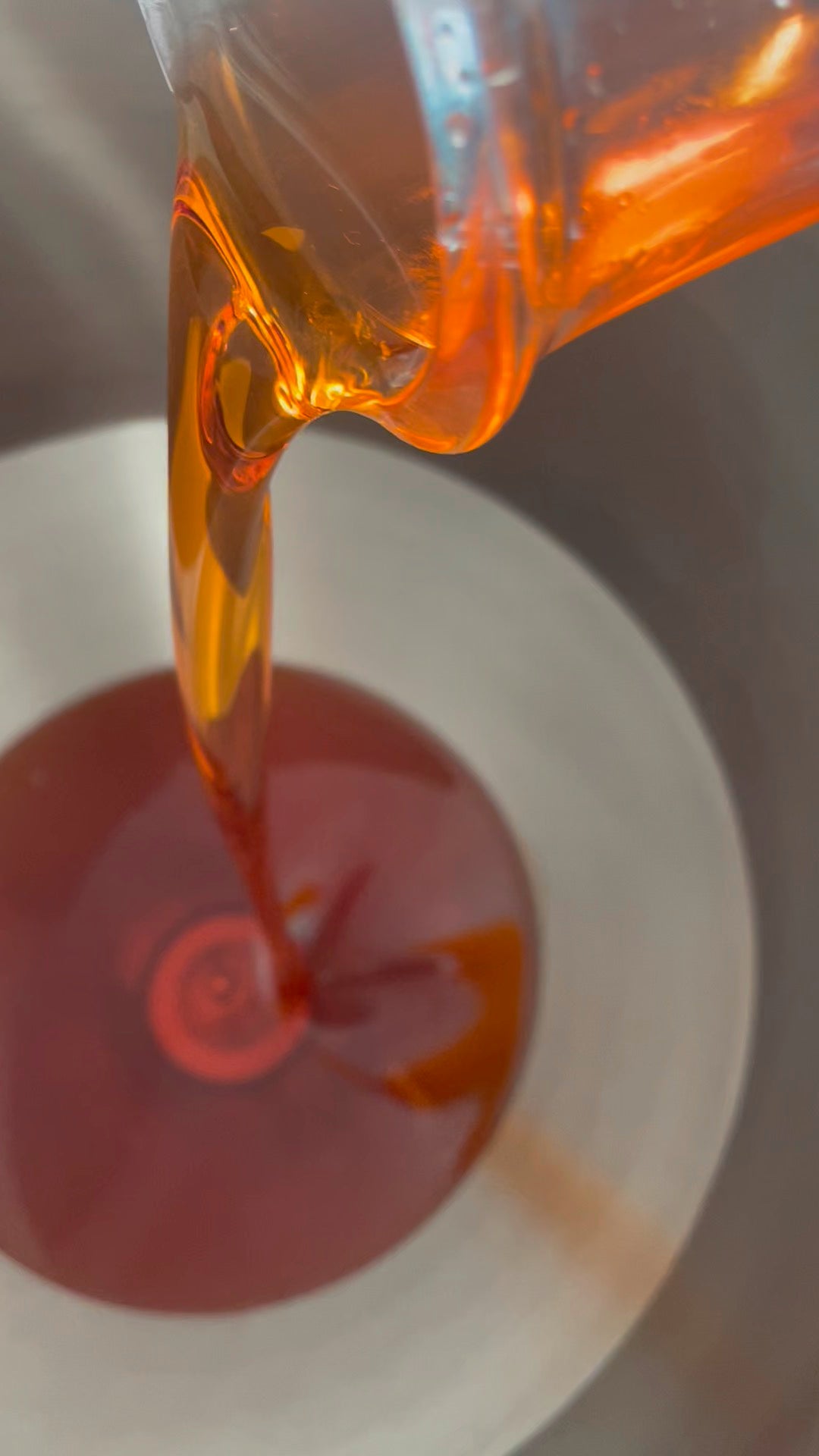 A close-up of Super Natural Beauty Oil, a translucent amber-orange liquid being poured from a clear container into a bottle filling machine.